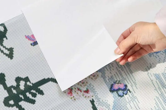 Hand verwijdert wit beschermvel van een onafgewerkte diamond painting met bloem- en symboolpatroon.