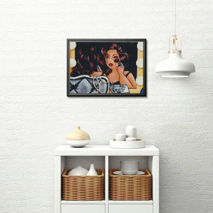 Ingelijst diamond painting van een vrouw die zich opmaakt voor een spiegel, opgehangen boven een witte kast met rieten manden en decoratieve objecten.